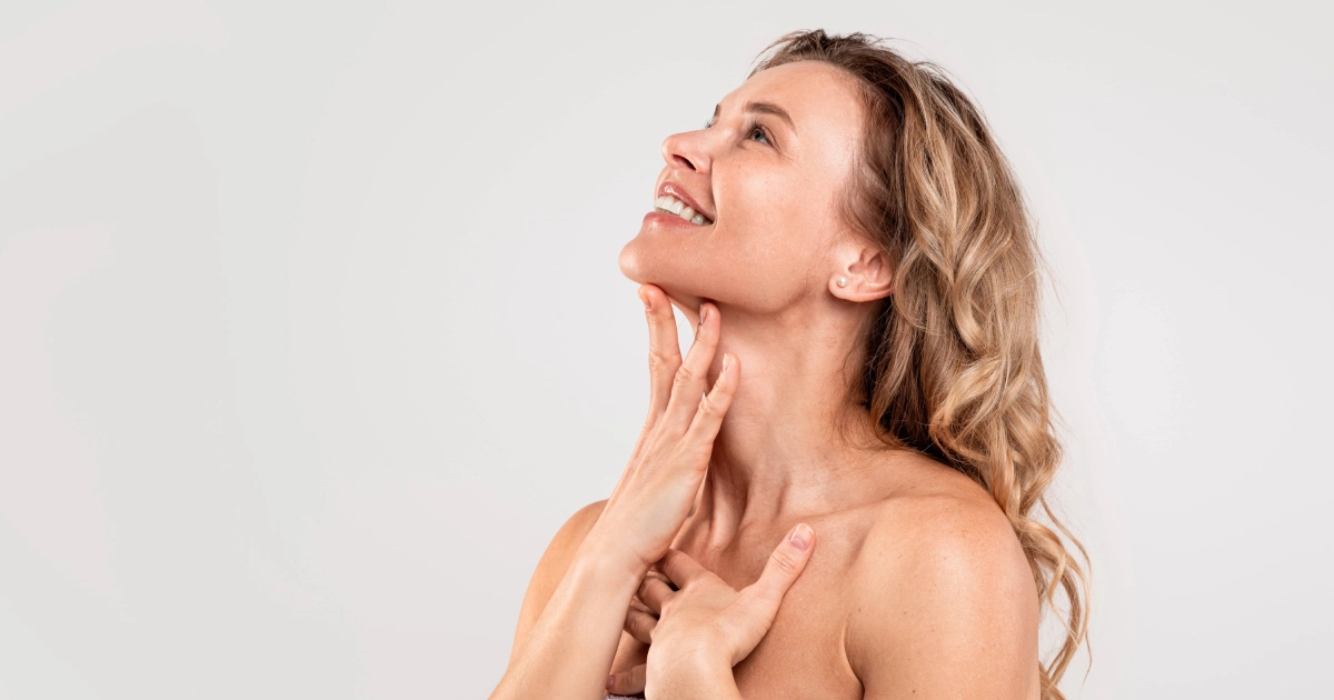A smiling middle-aged woman in a towel touches her neck and chest, looking up against a light background, highlighting Mesotherapy in Lake Charles, LA.